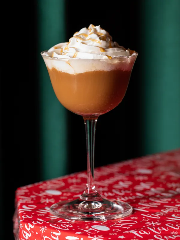 A glass of creamy cocktail with whipped cream and caramel 
                drizzle, set on a red patterned surface, captures the festive spirit at 
                Project Gigglewater in Toronto.