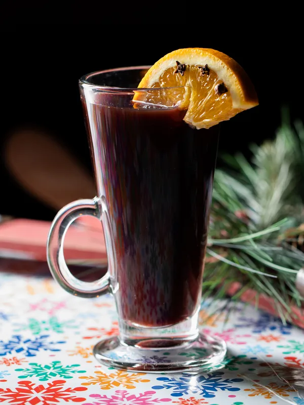 A glass of mulled wine, garnished with an orange slice and 
                cloves, sits invitingly on a colorful snowflake-patterned tablecloth at 
                Toronto's renowned Christmas Bar, Project Gigglewater. Pine branches add a 
                touch of festive elegance in the background.