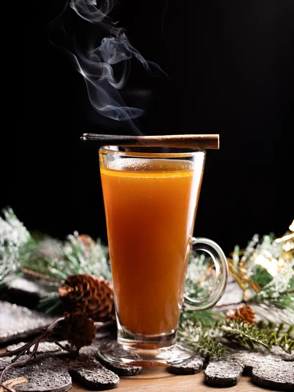 A steaming glass of hot cider with a cinnamon stick, 
              set on a wooden surface at Toronto's Christmas Bar, 
              Project Gigglewater, surrounded by pinecones and greenery.