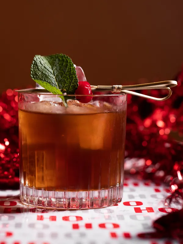 A glass of cocktail with ice, garnished with a mint sprig 
              and cherry on a skewer, sits elegantly at Project Gigglewater in 
              Toronto, reflecting the festive spirit with red decorations in the 
              background.