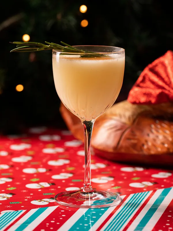 A frothy cocktail in a tall glass garnished with rosemary, 
                set on a festive red tablecloth with holiday patterns at Toronto's 
                Project Gigglewater.