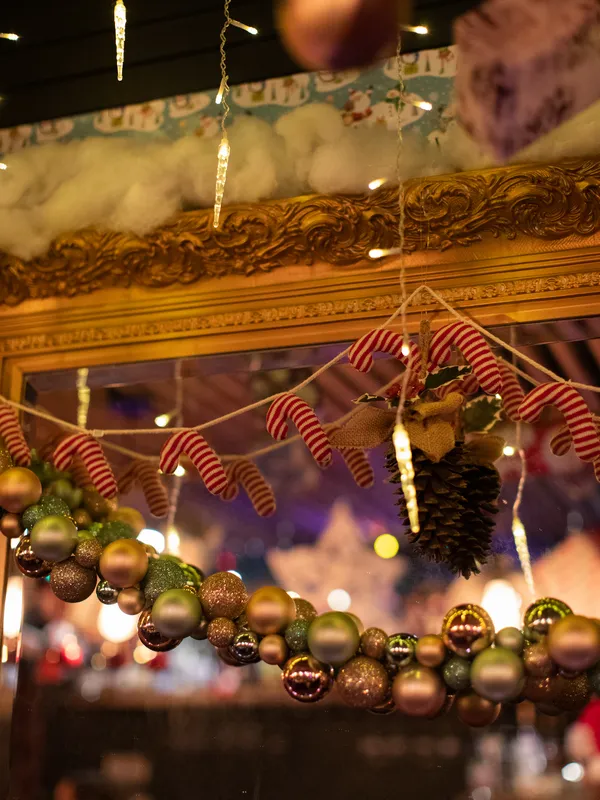 Christmas decorations featuring a pinecone, candy cane garland, 
          and ornaments hang on a mirror with a gold frame under icicle lights, creating 
          the perfect festive ambiance at Toronto's Christmas Bar in Project Gigglewater.