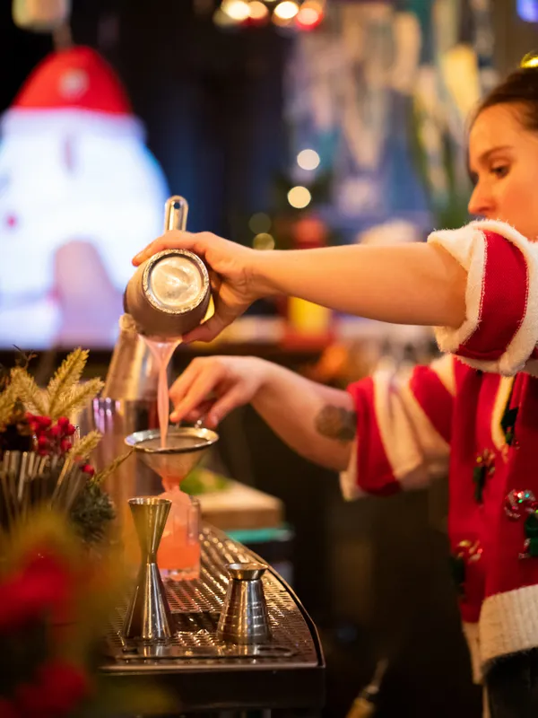 At Project Gigglewater in Toronto, a person in a festive sweater 
          pours a cocktail through a strainer at the Christmas bar, surrounded by 
          holiday decor.