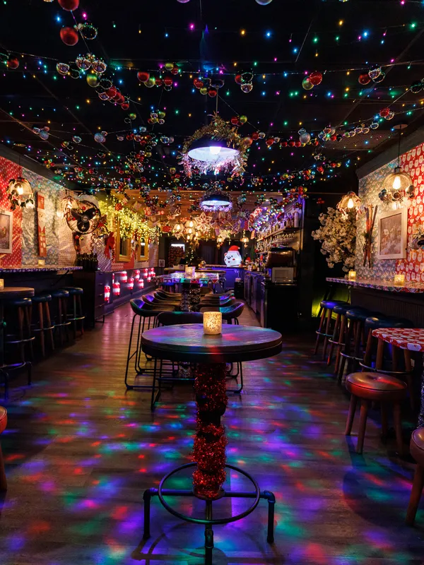Project Gigglewater in Toronto offers a festive bar interior 
            with colorful string lights, a decorated ceiling, and a central round table 
            adorned with a candle. The vibrant Christmas atmosphere creates enchanting 
            multicolored reflections on the floor, perfect for holiday gatherings.