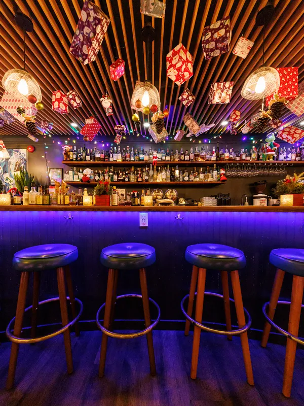 The vibrant interior of Project Gigglewater in Toronto features 
            a wooden ceiling adorned with colorful lights and festive decorations. 
            This Christmas bar is fully stocked with bottles, and four stools are 
            cozily arranged in front of the counter, inviting guests to celebrate the 
            season.
