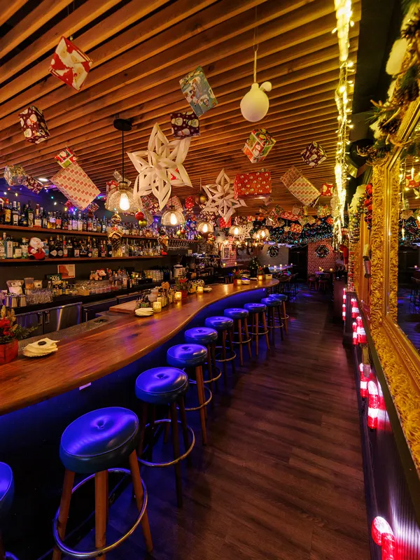 Project Gigglewater, a Christmas bar in Toronto, 
            features a festively decorated space with a wooden ceiling adorned with 
            hanging ornaments and a glowing countertop. Bar stools line the counter 
            while shelves brimming with bottles create an inviting backdrop.